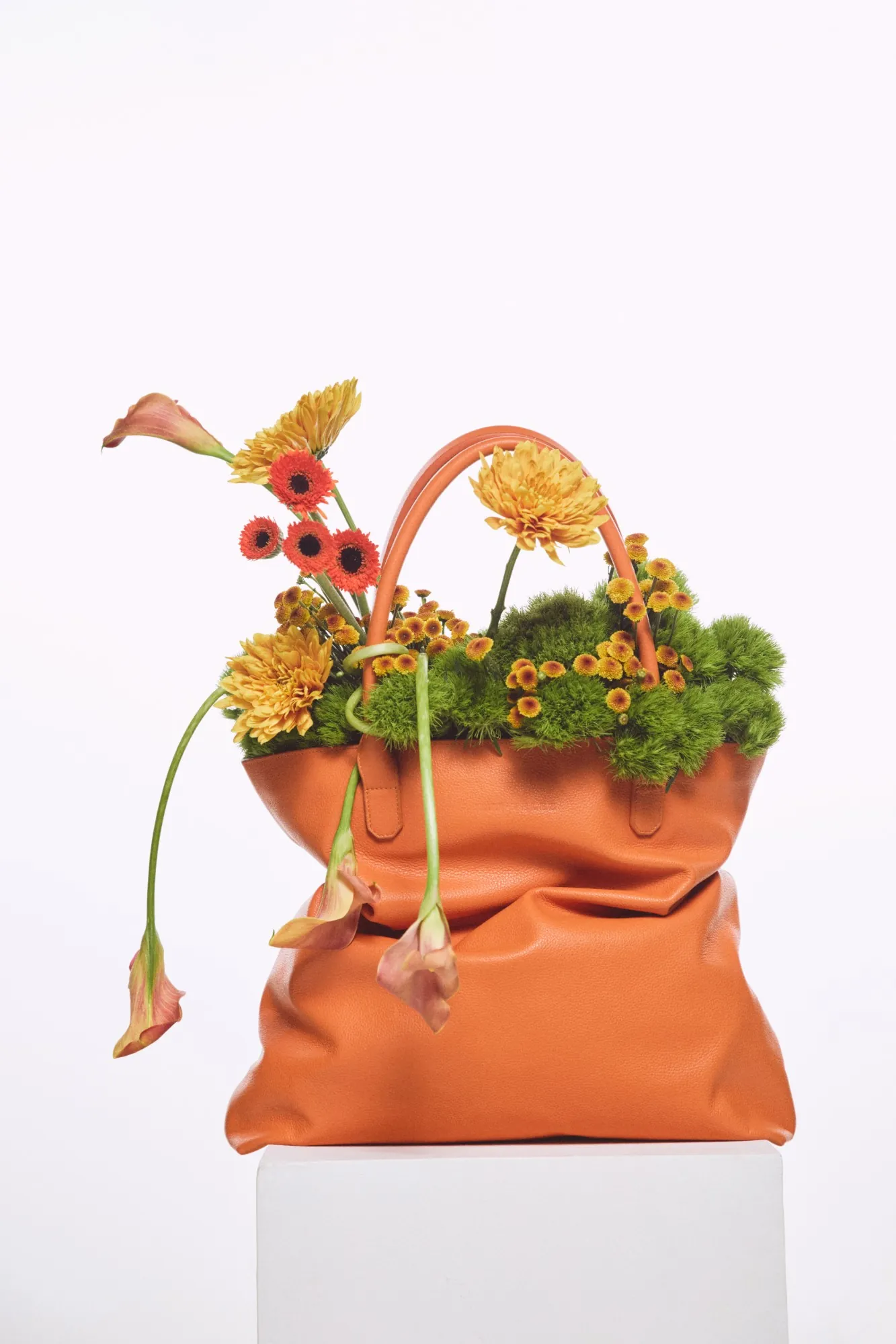 Bolso de cuero naranja de Vélez lleno de dianthus verde, botones de crisantemo, gerberas y calas, sobre un pedestal blanco